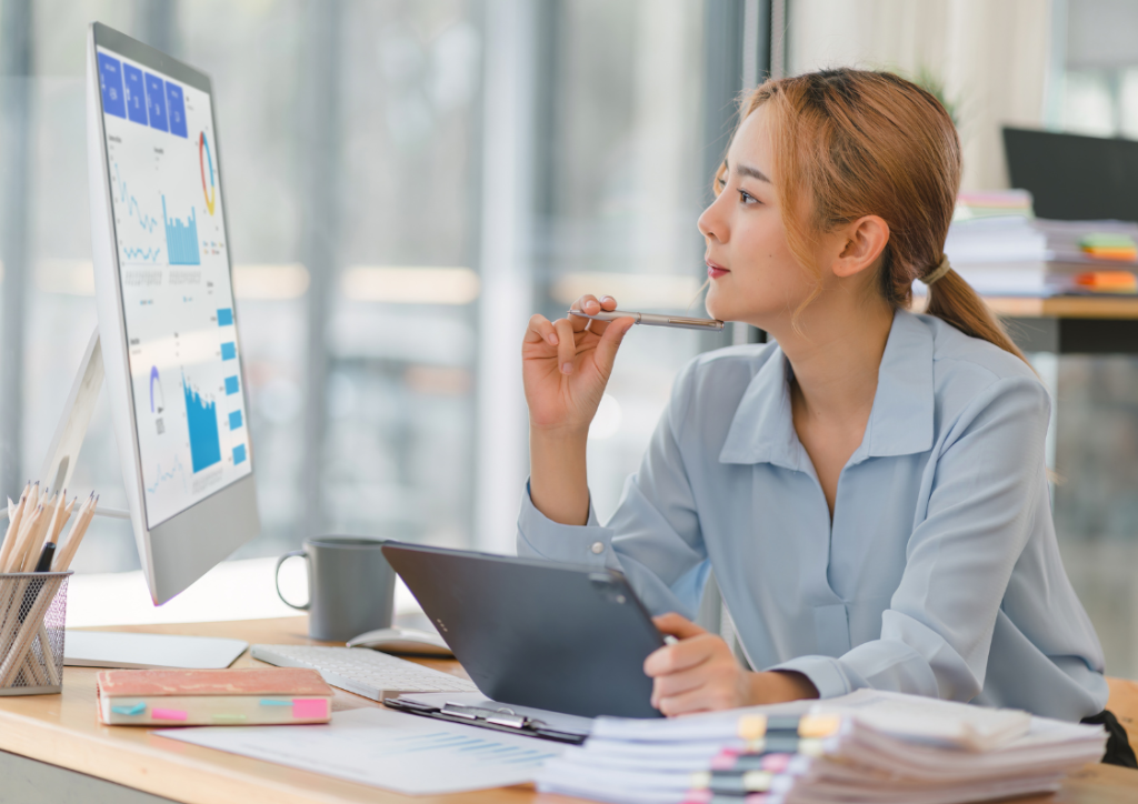 Business analyst examining data visualization dashboards on computer screen while holding a tablet, demonstrating modern business intelligence analysis at work. Professional woman in business attire analyzing BI reports and performance metrics in a corporate office setting.