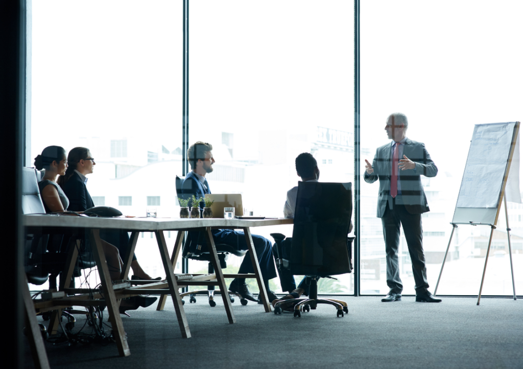 Business executives in a modern meeting room attending a presentation about IT cost optimization. A speaker stands by a flip chart while team members sit at a conference table near floor-to-ceiling windows.